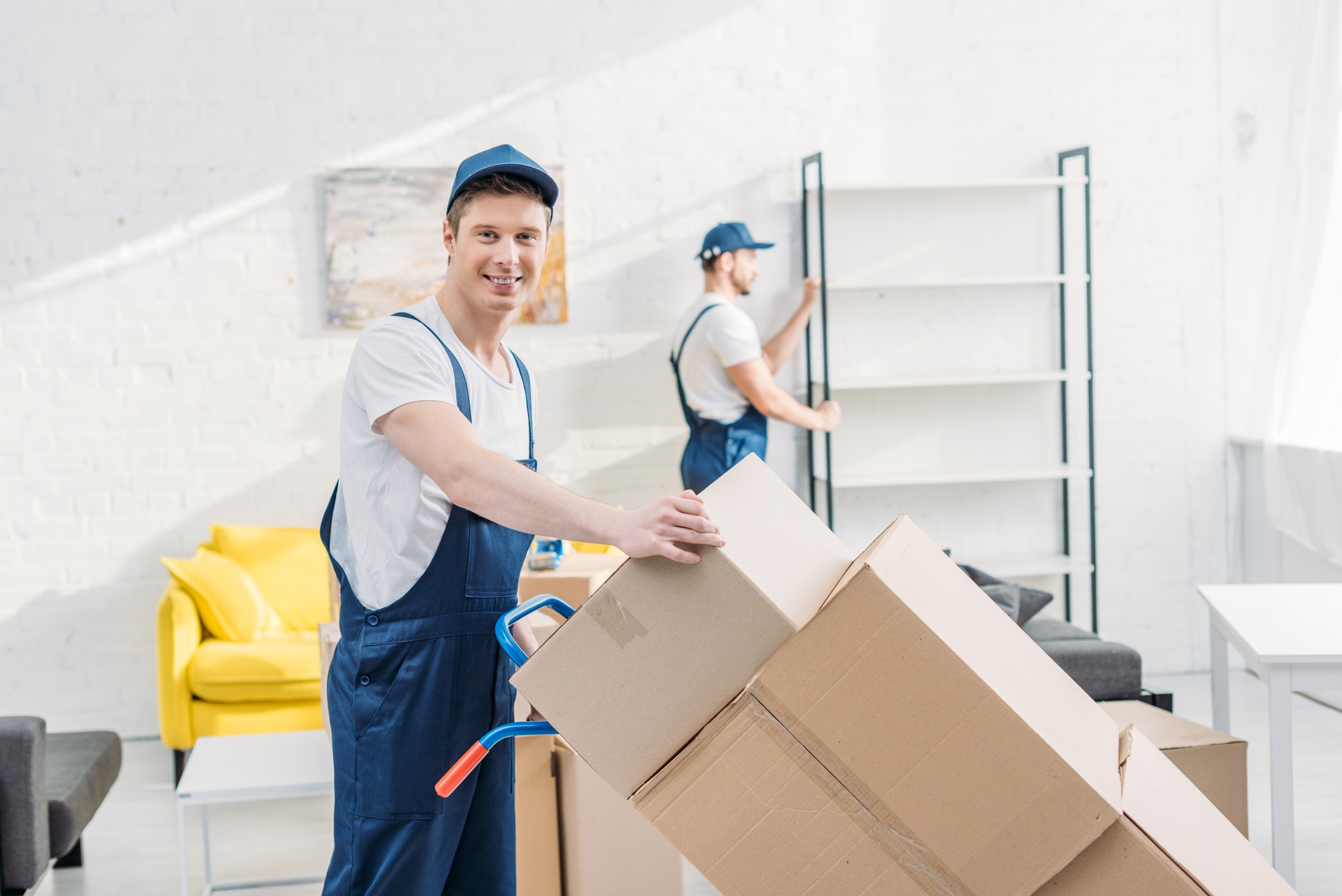 two movers in uniform transporting cardboard boxes and furniture in apartment